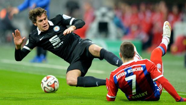 Ein Fußballspieler in Schwarz grätscht nach dem Ball, während ein Spieler von Bayern München zu Boden geht.
