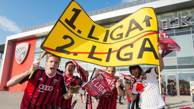 Fußballfans des FC Ingolstadt feiern mit einem Banner „1. Liga, 2. Liga“ vor dem Stadion.