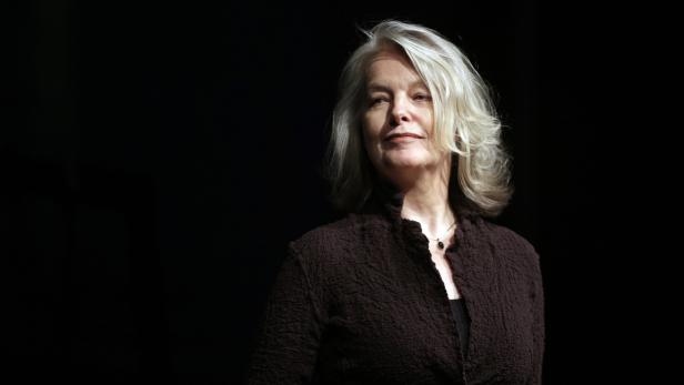 Eine Frau mit grauen Haaren und brauner Jacke vor schwarzem Hintergrund.