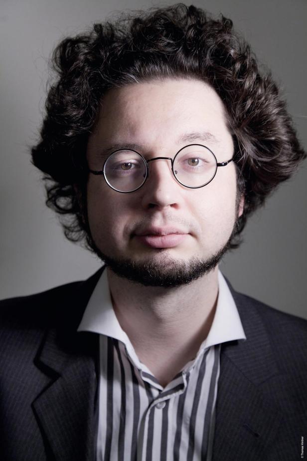 Ein Mann mit lockigem Haar, Brille und Bart trägt einen Anzug.