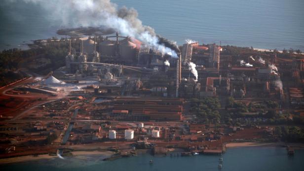 Luftaufnahme einer großen Industrieanlage an der Küste, aus der Rauch aufsteigt.