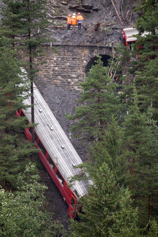 Ein entgleister roter Zug liegt an einem Hang neben einem Tunnel. Drei Arbeiter stehen oberhalb des Tunnels.