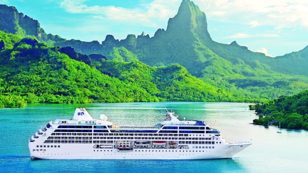 Ein Kreuzfahrtschiff fährt vor einer tropischen Insel mit üppiger Vegetation.