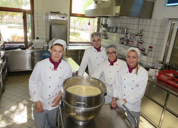 Vier junge Köche stehen in einer Küche um einen großen Topf mit Teig.