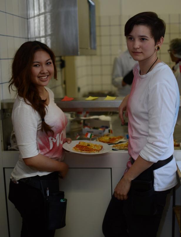 Zwei junge Frauen stehen in einer Küche, eine hält einen Teller mit Pizza.