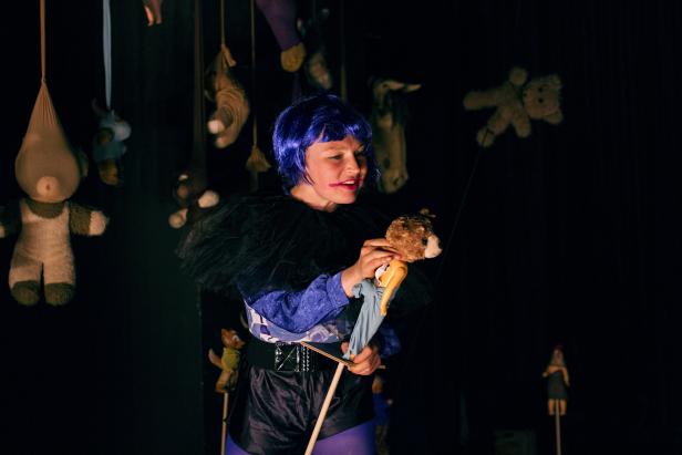 Eine junge Frau mit blauer Perücke spielt mit einer Teddybär-Marionette vor einem dunklen Hintergrund mit Stofftieren.