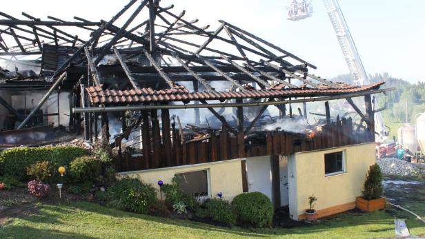 Ein Haus ist nach einem Brand stark beschädigt, mit Feuerwehrleuten im Hintergrund.