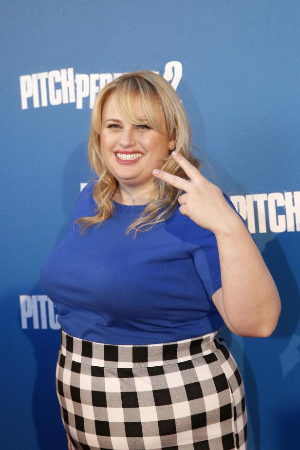 Rebel Wilson lächelt und zeigt das Victory-Zeichen vor dem Hintergrund von „Pitch Perfect 2“.