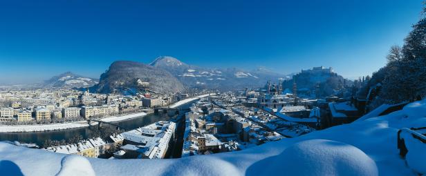 Winterliche Panoramaansicht von Salzburg mit schneebedeckten Dächern und Bergen.