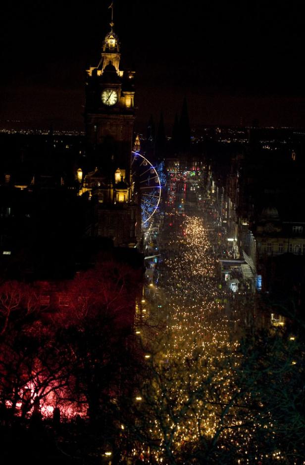 Nachtsicht auf Edinburgh mit Menschen, die eine von Lichtern gesäumte Straße entlanggehen, und einem beleuchteten Riesenrad.