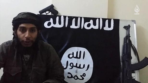 Ein Mann mit Turban vor einer Flagge mit islamischen Schriftzeichen, einer Pistole und einem Gewehr.