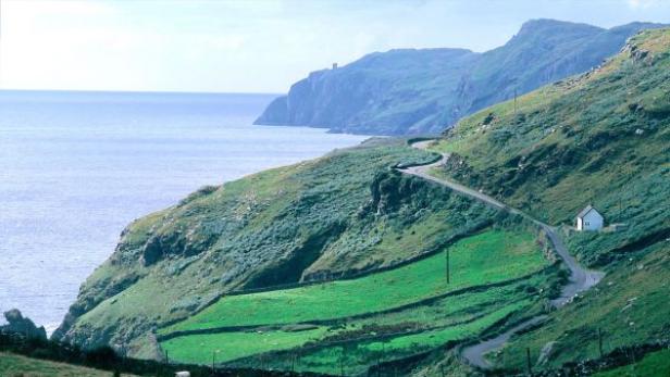 Eine kurvenreiche Straße führt an der Küste entlang, vorbei an grünen Hügeln und einem kleinen weißen Haus.