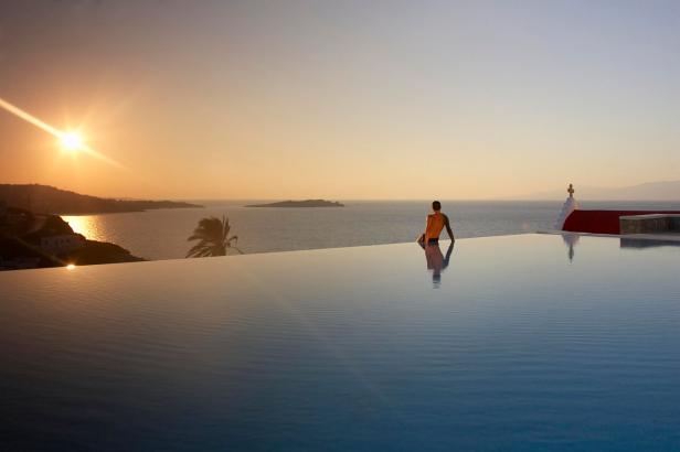 Ein Mann sitzt am Rand eines Infinity-Pools und blickt auf das Meer bei Sonnenuntergang.