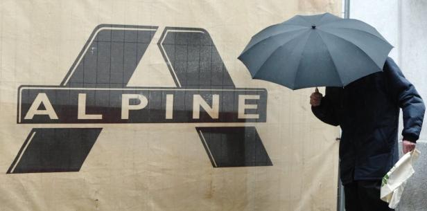 Eine Person mit Regenschirm steht vor einem Plakat mit dem Schriftzug „Alpine“.