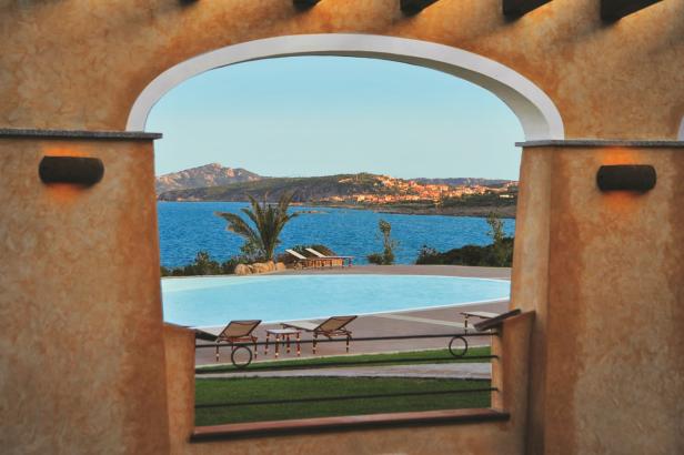 Blick durch einen Torbogen auf einen Pool und das Meer mit einer Küstenstadt im Hintergrund.