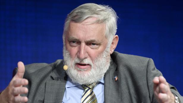 Ein Mann mit grauem Haar und Bart gestikuliert vor einem blauen Hintergrund.