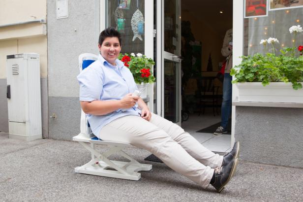 Eine Frau sitzt auf einem Treppenlift vor einem Geschäft und lächelt.