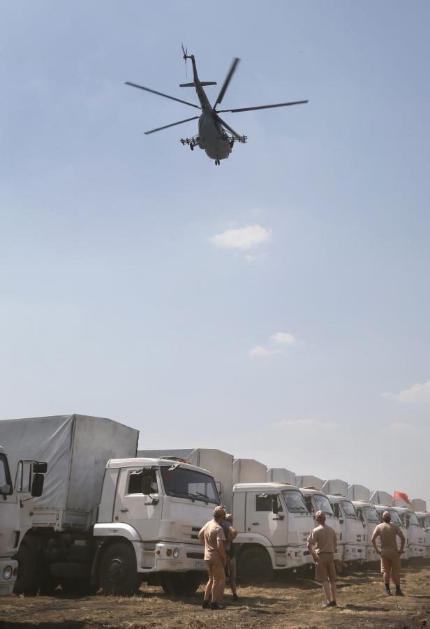 Ein bewaffneter Hubschrauber überfliegt eine Reihe weißer Militärlastwagen.