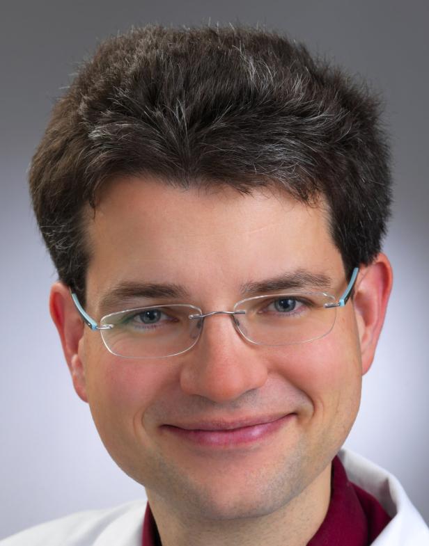 Ein lächelnder Mann mit Brille und kurzem, dunklem Haar vor einem grauen Hintergrund.