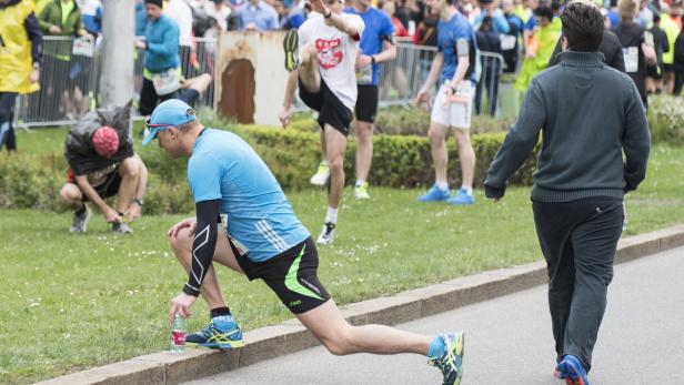 Ein Läufer dehnt sich am Straßenrand, während andere Läufer im Hintergrund zu sehen sind.