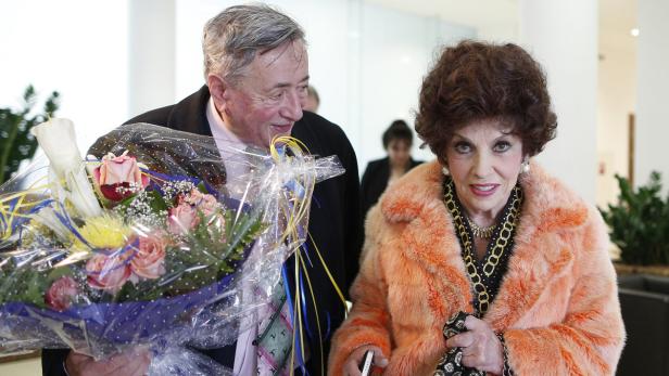 Eine ältere Frau im Pelzmantel und ein Mann mit einem Blumenstrauß.