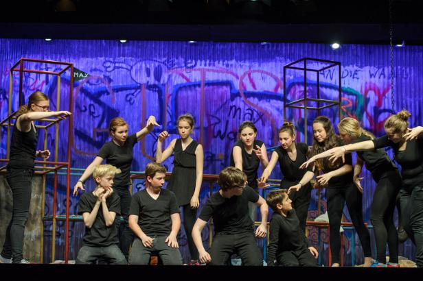Eine Gruppe von Jugendlichen in schwarzen Kostümen steht auf einer Bühne vor einer Graffiti-Wand.