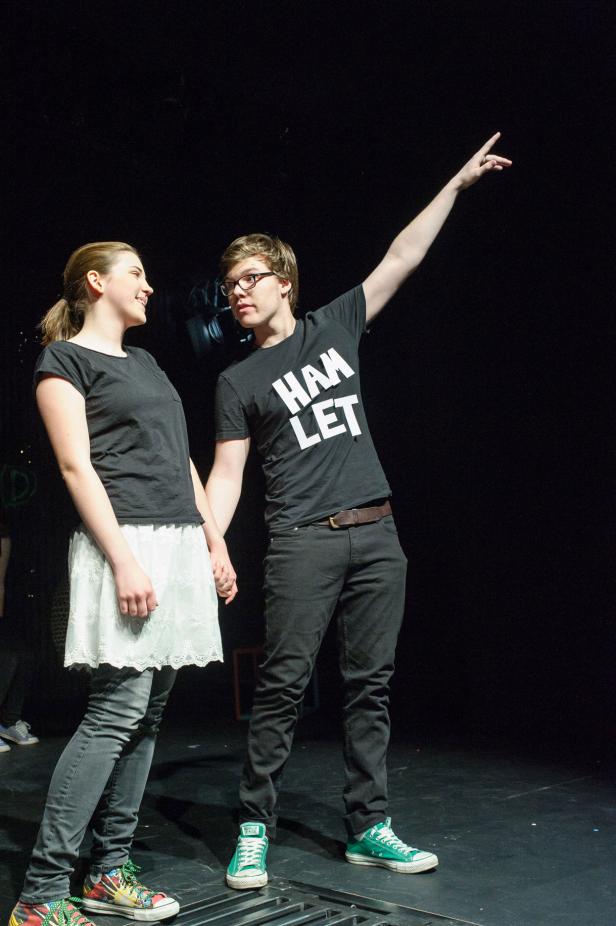 Zwei Schauspieler stehen auf der Bühne, einer mit einem „Hamlet“-T-Shirt, und halten Händchen.