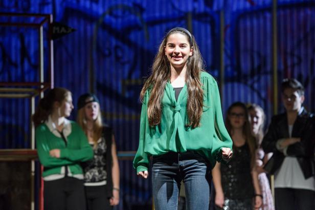 Eine lächelnde junge Frau in einem grünen Oberteil steht auf einer Bühne vor anderen Darstellern.