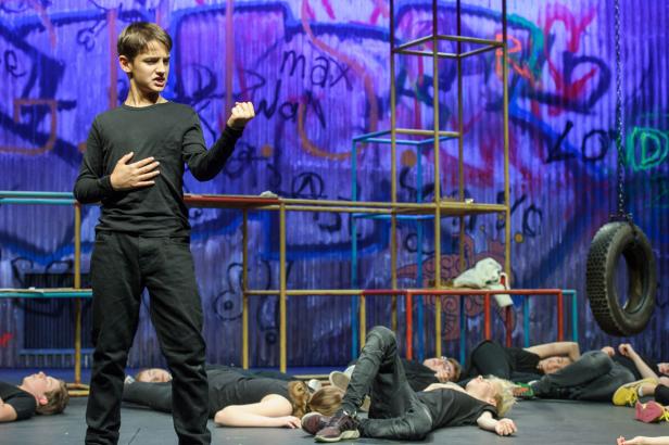 Ein junger Schauspieler steht vor einer Gruppe liegender Personen auf einer Bühne mit Graffiti-Hintergrund.
