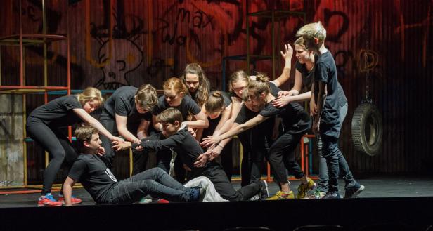 Eine Gruppe von Jugendlichen umringt auf einer Bühne einen am Boden liegenden Jungen.