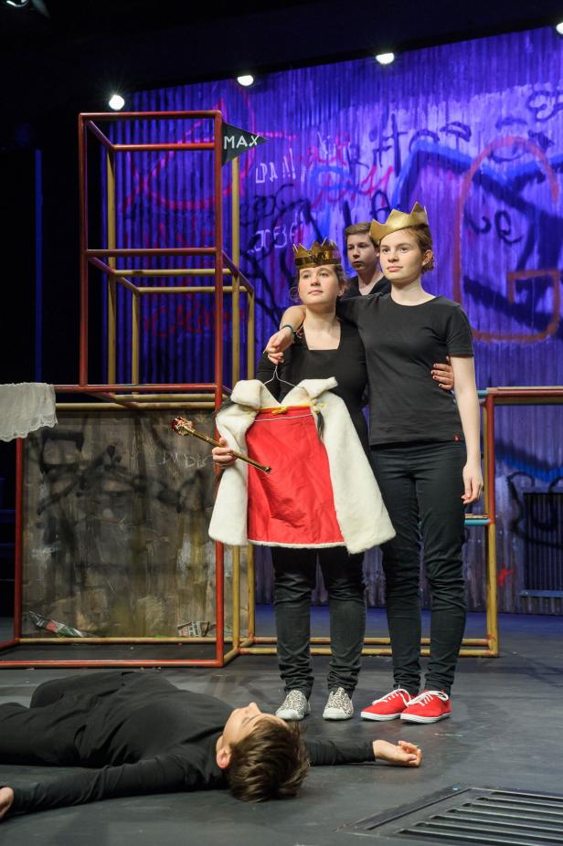 Drei Jugendliche mit Kronen auf einer Bühne, einer liegt am Boden.