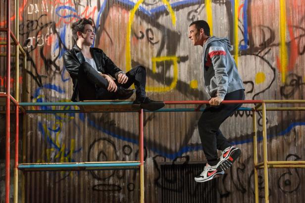 Zwei junge Männer sitzen und stehen auf einem Spielgerüst vor einer Graffitiwand.