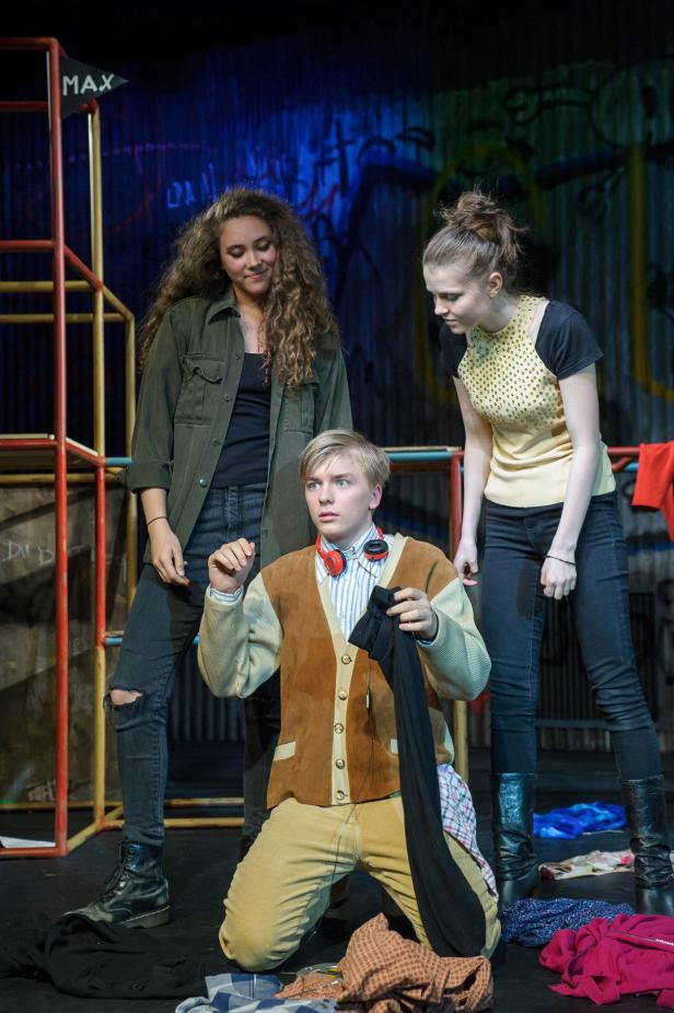 Drei junge Schauspieler auf einer Bühne mit Kleidern und einer Leiter im Hintergrund.