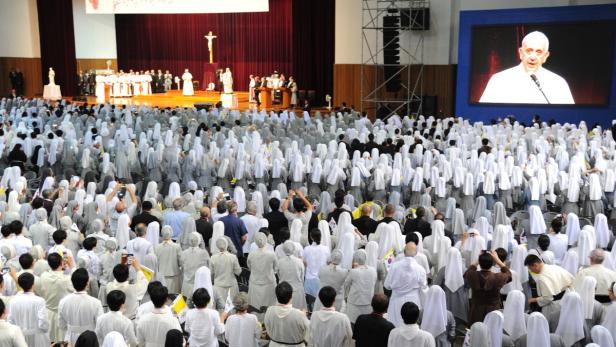 Eine große Menschenmenge, darunter viele Nonnen, versammelt sich vor einer Bühne, auf der Papst Franziskus auf einem Bildschirm zu sehen ist.
