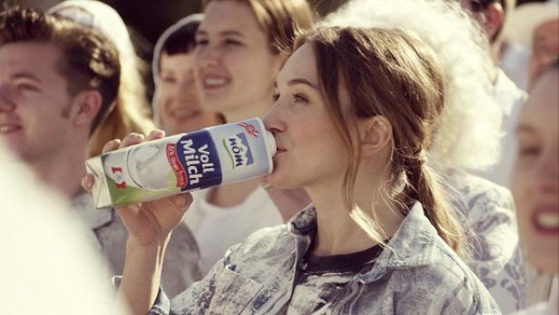 NÖM - Markenkampagne 2015 - TV-Spot Bild 12 Eine junge Frau trinkt Vollmilch aus einem Tetrapack vor einer Gruppe von Menschen.