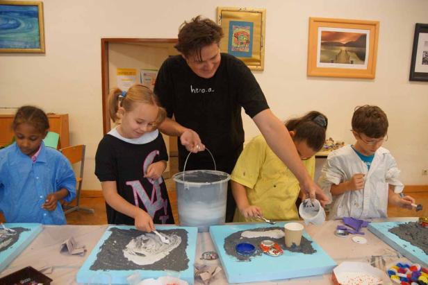 Eine Gruppe Kinder gestaltet unter Aufsicht Reliefs mit Farbe und verschiedenen Materialien.