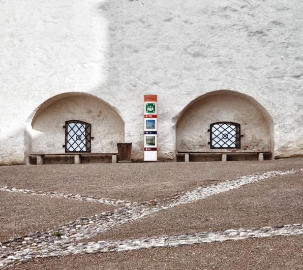 Eine Informationstafel steht zwischen zwei vergitterten Fenstern in einer weißen Steinmauer.