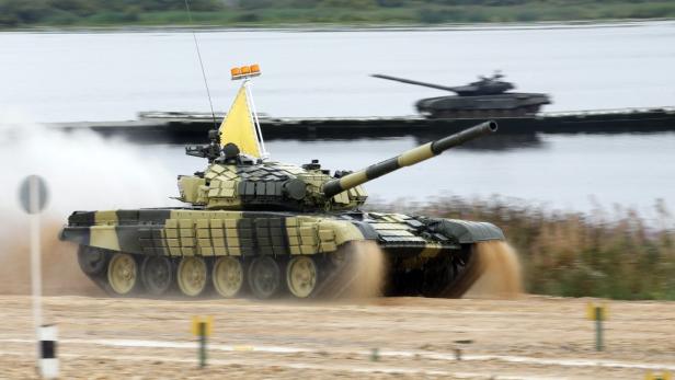 Ein olivgrüner Panzer fährt über eine Sandpiste, im Hintergrund ein Gewässer.