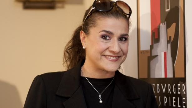 Eine lächelnde Frau mit Sonnenbrille im Haar und einer Halskette mit Perlenanhänger.