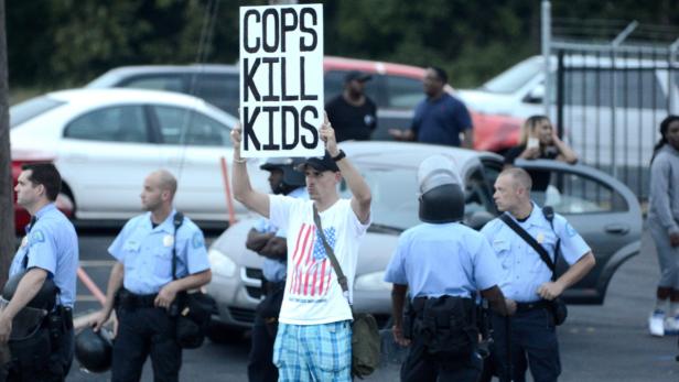 Ein Demonstrant hält ein Schild mit der Aufschrift „Cops Kill Kids“ inmitten einer Gruppe von Polizisten.