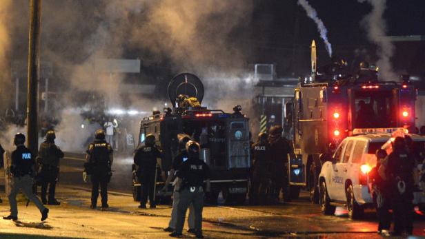 Polizisten stehen in der Nacht vor gepanzerten Fahrzeugen im Rauch.