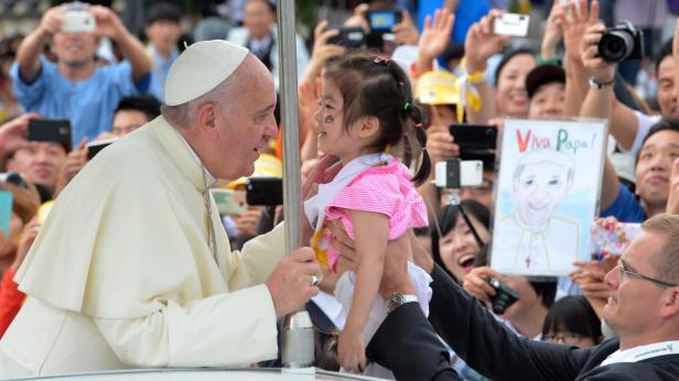 Papst Franziskus hält ein kleines Mädchen in seinen Armen, umringt von einer Menschenmenge.