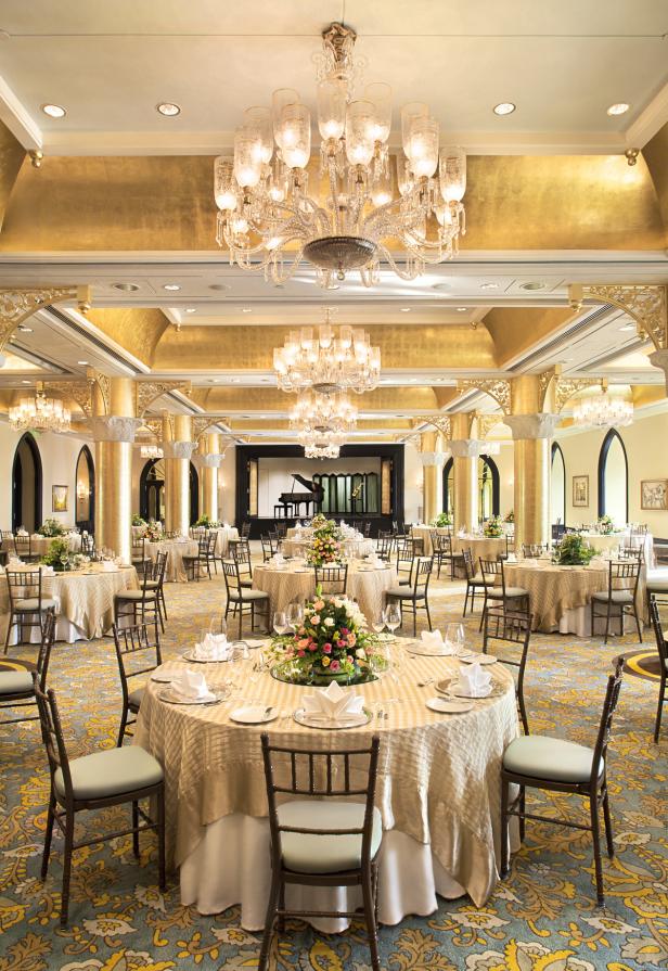 Ein festlicher Ballsaal mit runden Tischen, Blumengestecken und einem Klavier auf der Bühne.