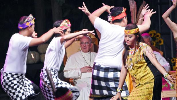 Papst Franziskus sitzt auf einem Stuhl, während Tänzer in traditionellen Kostümen vor ihm auftreten.