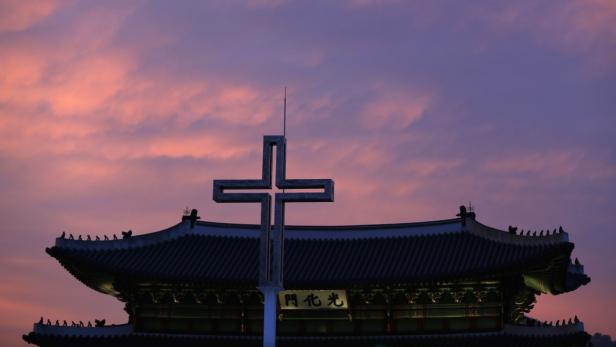 Ein Kreuz steht vor einem traditionellen koreanischen Gebäude bei Sonnenuntergang.