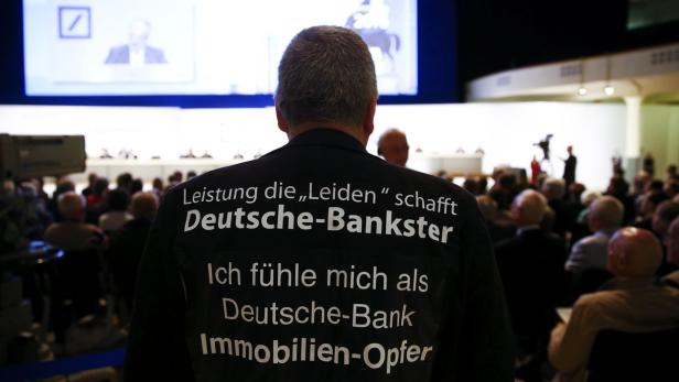 Ein Mann mit einem T-Shirt mit der Aufschrift „Deutsche Bank Immobilien-Opfer“ steht in einem Publikum.