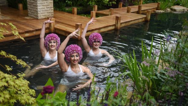 Drei Frauen mit Blumenkopfbedeckungen stehen in einem Teich mit einem Holzsteg im Hintergrund.