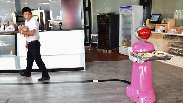 Ein rosafarbener Roboter serviert Essen, während ein Kellner im Hintergrund vorbeigeht.