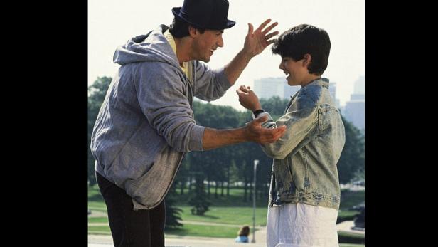 Sylvester Stallone mit Hut interagiert mit einem Kind in einer Parksituation.