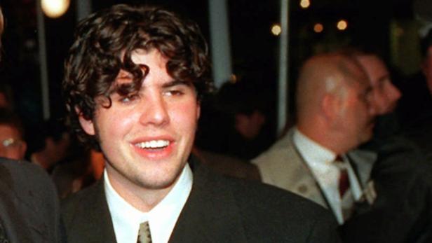 Ein junger Mann mit lockigem Haar lächelt in einem dunklen Anzug.